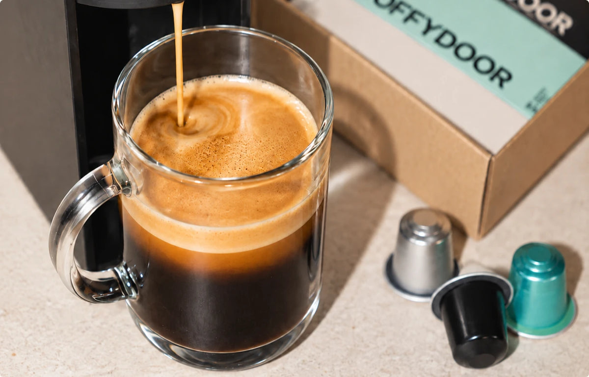 Coffee pouring into a glass mug beside a Coffydoor box and three pods