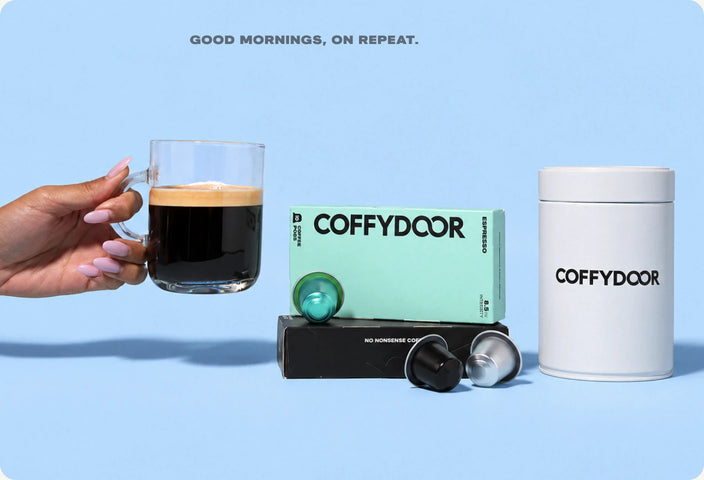 A hand holding a glass mug of Coffydoor coffee with thick crema, beside a pack of Coffydoor espresso pods and a Coffydoor tin. Good mornings, on repeat.